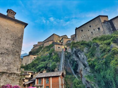 Önünde pembe çiçekler olan pitoresk bir dağ vadisinde canlı bir günbatımı, tuhaf bir köyden geçen bir yol, ve arka planda dramatik uçurumlar.