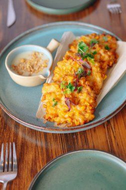 Masada bir tabak kızarmış mısır, mavi tabak ve beyaz kase. Mısır maydanozla süslenir ve sosla servis edilir.