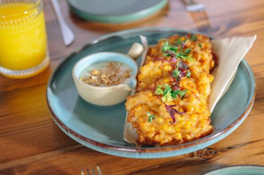 Tahta bir masada bir tabak kızarmış mısır ve bir bardak portakal suyu. Mısır sosuyla servis edilir ve maydanozla süslenir.