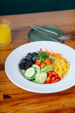 Domates, salatalık ve havuç gibi çeşitli sebzelerle dolu renkli bir salata. Masada salatanın yanında bir bardak portakal suyu var.