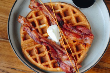 Bir tabak domuz pastırması ve şurup serpiştirilmiş waffle. Tabak ahşap bir masanın üzerinde.