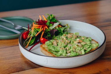 Bir tabak avokado tostu ve yanında salata. Tabak ahşap bir masanın üzerinde. Salata marul, havuç ve domateslerden oluşur.