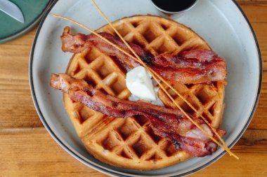 Ortasında çubuk olan bir tabak pastırma ve waffle. Pastırma pişti ve waffle 'lar altın sarısı. Tabak ahşap bir masanın üzerinde.