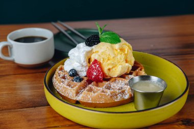 Krem şantili ve çilekli waffle tabağı bir fincan kahvenin yanında duruyor.