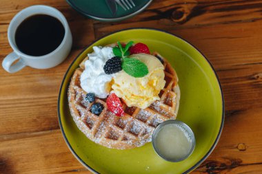 Krem şantili ve meyveli bir tabak waffle bir fincan kahvenin yanında duruyor.