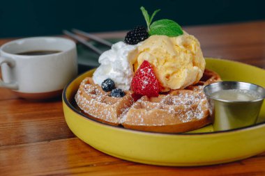 Krem şantili ve çilekli waffle tabağı bir fincan kahvenin yanında duruyor.