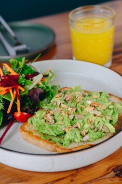 Bir tabak avokado tostu, yanında salata ve bir bardak portakal suyu. Tabak ahşap bir masaya konmuş.