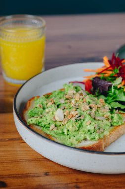 Bir tabak avokado tostu, yanında sebze ve bir bardak portakal suyu. Tabak ahşap bir masaya konmuş.