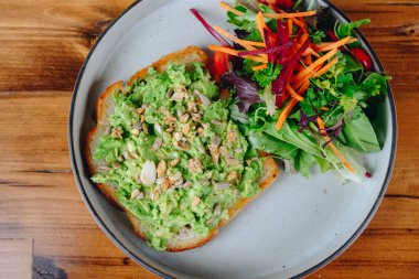 Bir tabak yemek, sandviç ve salata. Sandviç avokadodan yapılır ve üzerinde fındık vardır. Salata marul, havuç ve diğer sebzelerden yapılır. Tabak ahşap bir masanın üzerinde.