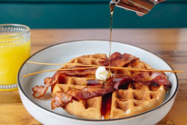 Üzerine şurup dökülmüş bir tabak pastırma ve waffle. Masada bir bardak portakal suyu var.