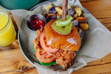 Hamburger ve yanında patates olan bir tabak yemek. Hamburgerin üstünde turşu ve domates var. Tabak ahşap bir masanın üzerinde.