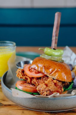 Ekmeğin üstünde hamburger ve yeşil biberli bir tabak yemek. Masada bir bardak portakal suyu var.