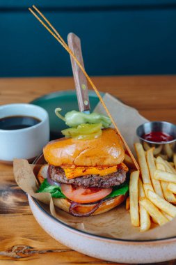 Bıçak saplanmış bir hamburger ve yanında patates kızartması. Hamburger bir dilim turşu ve bir domatesle kaplıdır. Masa düzeninde bir fincan kahve ve ketçap şişesi var.