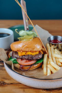 Üstünde turşu mızrağı ve yanında patates kızartması olan bir hamburger. Hamburger, kahverengi bir peçeteyle beyaz bir tabakta servis edilir. Görüntü gelişigüzel ve rahat bir atmosfer sağlıyor.