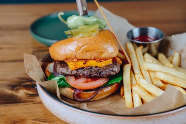 Üstünde turşu mızrağı ve yanında patates kızartması olan bir hamburger. Hamburger tabakta, altında peçete var.