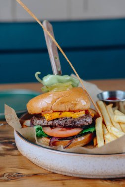 Üstünde turşu mızrağı ve yanında patates kızartması olan bir hamburger. Hamburger beyaz bir tabakta servis ediliyor ve patates kızartmaları da kağıt bir sepette.