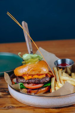 Üstünde turşu mızrağı ve yanında patates kızartması olan bir hamburger. Hamburger beyaz tabakta servis ediliyor ve patatesler küçük bir kasede. Sahne rahat ve rahat.