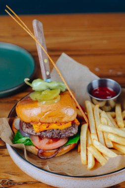 Üstünde turşu mızrağı ve yanında patates kızartması olan bir hamburger. Hamburger beyaz tabakta, kahverengi peçeteyle servis edilir.