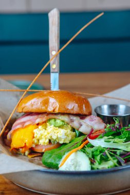 Pastırmalı, yumurtalı ve avokadolu bir sandviç bir tabakta salatanın yanında duruyor. Sandviçin üzerinde bir bıçak ve bir parça brokoli var. Salata havuç ve kerevizle süslenmiş.