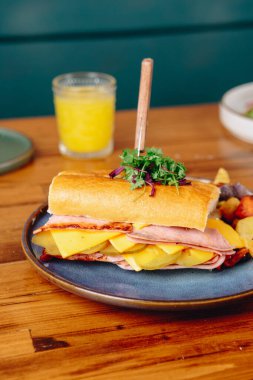 Bir bardak portakal suyu ve mavi tabakta jambonlu ve peynirli sandviç. Sandviçin üzerinde maydanoz ve kürdan var.
