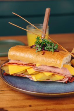 Jambonlu, peynirli ve turşulu bir sandviç mavi tabakta duruyor. Sandviçin yanında bir bardak portakal suyu var.