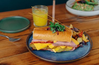 Mavi tabakta jambonlu ve peynirli bir sandviç ahşap bir masada oturur. Sandviçin yanında bir bardak portakal suyu var.