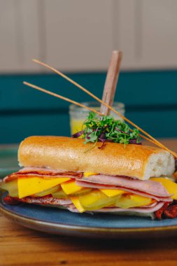 Mavi tabakta jambonlu, peynirli ve turşulu bir sandviç. Sandviç ikiye bölünmüş ve üstünde yeşil bir garnitür var.
