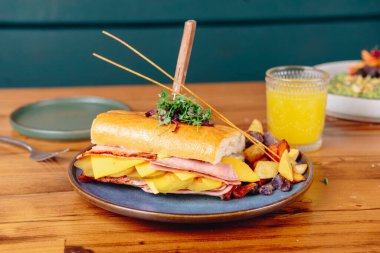 Üzerinde kürdan olan bir sandviç mavi bir tabakta servis edilir. Tabak ahşap bir masanın üzerine konur. Ayrıca masada havuç ve bir bardak portakal suyu var.