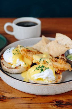 Bir tabak cips ve bir fincan kahveyle birlikte Benedict usulü yumurta. Tabak ahşap bir masaya konmuş.