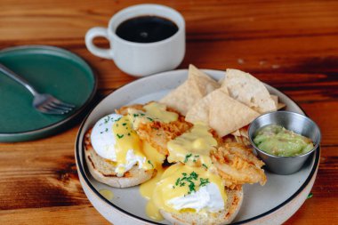 Yanında cips ve guacamole ile bir tabak kahvaltı yemeği. Tabak arka planda bir fincan kahveyle ahşap bir masanın üzerine kurulmuştur. Sahne rahat ve rahat.