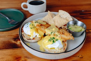 Bir tabak cips ve guacamole soslu Benedict yumurtası. Tabak arka planda bir fincan kahveyle ahşap bir masanın üzerine kurulmuştur. Sahne sıradan ve davetkar.