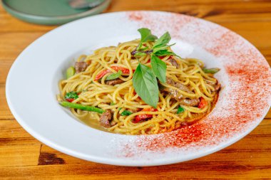 Bir tabak etli ve sebzeli erişte. Erişte kırmızı ve beyaz ve et kahverengidir. Tabak ahşap bir masanın üzerinde.