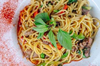 Bir tabak etli ve sebzeli erişte. Erişteler uzun ve sarı ve et kahverengi. Sebzeler yeşil ve kırmızı. Yemek lezzetli ve iştah açıcı görünüyor.