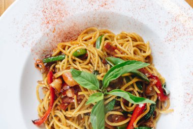 Sebzeli ve etli bir tabak erişte. Erişteler kırmızı biber taneleriyle kaplı. Tabak beyaz bir kasede servis edilir.