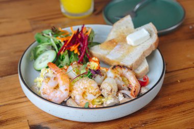 Karides, ekmek ve sebzeli bir tabak yemek. Tabak ahşap bir masanın üzerinde.