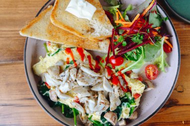 Bir tabak yemek, bir parça tost ve salata. Salatada havuç, domates ve marul gibi çeşitli sebzeler vardır. Tabak ahşap bir masaya konmuş.