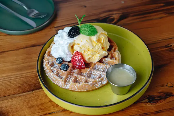 Yeşil tabağın üstünde dondurmalı ve meyveli waffle. Tabak ahşap bir masanın üzerinde.