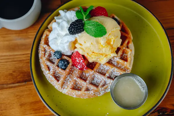 Üstünde krema ve böğürtlen olan bir tabak waffle. Tabak bir bardağın yanındaki masada.