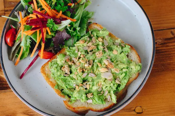 A plate of avocado toast with a side of salad. The avocado toast is topped with nuts and seeds, and the salad is made up of lettuce, carrots, and tomatoes. The plate is set on a wooden table