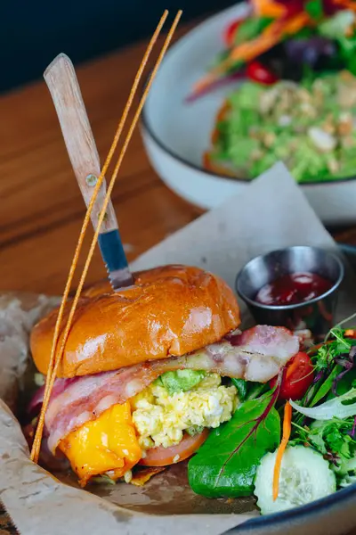 Bir pastırmalı ve yumurtalı sandviç, yanında sebze ile birlikte bir çörek üzerinde servis edilir. Sandviçin üzerinde pastırma, peynir ve domates var. Sebzeler arasında marul, salatalık ve havuç var.