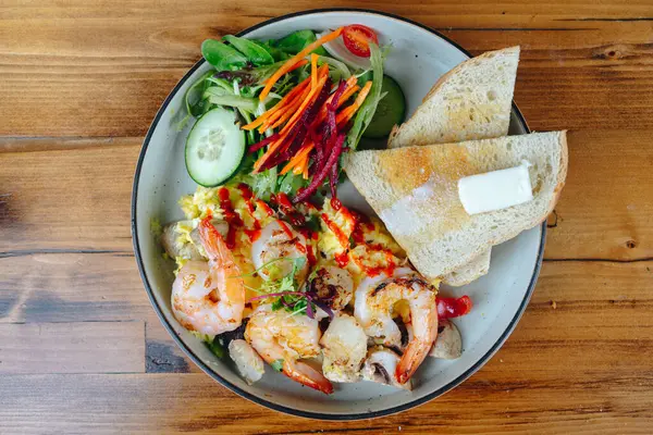 Bir tabak karides, sebze ve tost. Tabak ahşap bir masanın üzerinde.