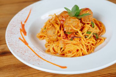 Köfteli bir tabak spagetti ve üstünde yeşil bir yaprak. Tabak ahşap bir masada servis edilir.