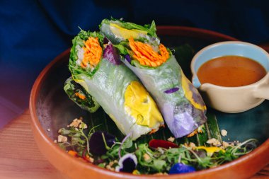 Bir tabak yemek, iki paket ve bir kase sos. Paketler sebzelerle dolu ve sos kremalı, kahverengi bir renk. Tabak ahşap bir masaya konmuş.