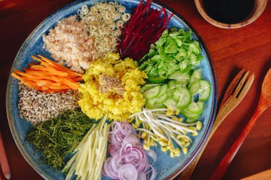 Havuç, salatalık ve kereviz de dahil olmak üzere çeşitli sebzelerle dolu mavi bir tabak. Tabak, yanında çatal ve kaşıkla ahşap bir masaya konmuş.