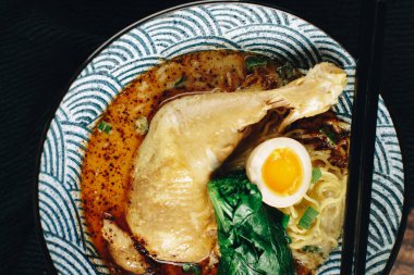 Üzerine bir parça tavuk ve yumurta olan bir kase çorba. Çorba zengin, kremsi bir renktir ve tavuk ve yumurta yemeğe renk ve doku katar. Sahne sıcak ve davetkar.