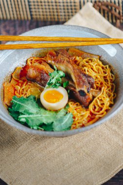 Bir kase etli ve sebzeli erişte ve üstüne de bir parça yumurta. Kase kahverengi bir bezle bir masanın üzerinde.