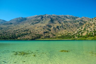 Yunanistan 'ın Girit kentinde mavi gökyüzü altında dağlık manzarayla birlikte Kournas Gölü' nün berrak turkuaz suları