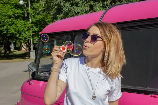 Beautiful girl in glasses and white T-shirt blows soap balls on the background of pink car