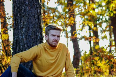 A bearded young man of European appearance is sitting on the ground in the autumn forest