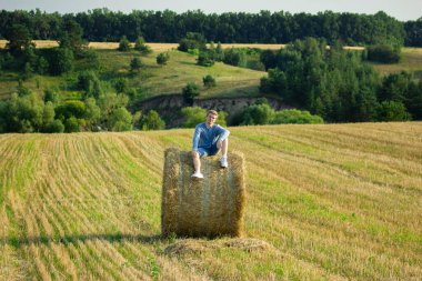 A guy sitting on a large bale of straw in an empty field of cut wheat